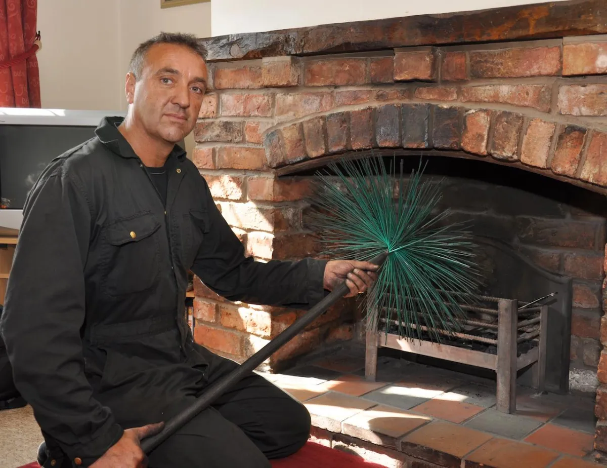 Rob Eckersley, professional chimney sweep, kneeling with a sweeping brush beside a brick inglenook fireplace